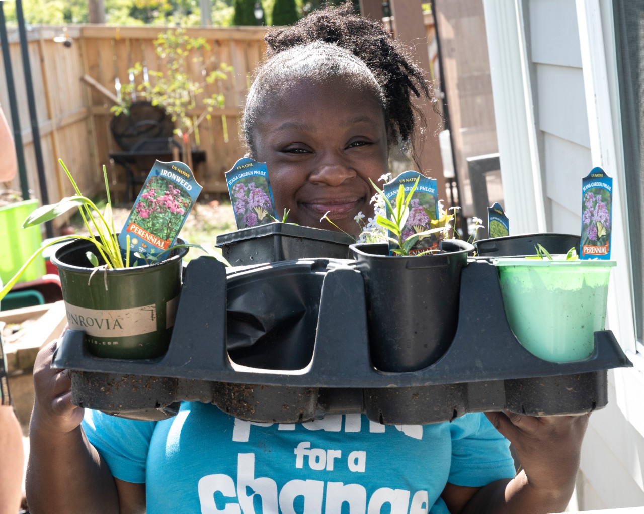 a young Black woman smiling very big, standing outside on a sunny day, and wearing a blue t-shirt that says “Plant for a Change.” She is holding up a tray of small potted plants just starting to sprout, with signs showing which plants or flowers they are, and smiling over the tray at the camera.