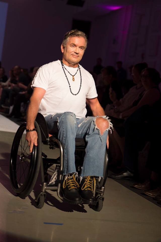 a man pushing a manual wheelchair through an audience. He is wearing an original fashion design, which includes a white t-shirt and ripped blue jeans. He is also wearing two necklaces. 