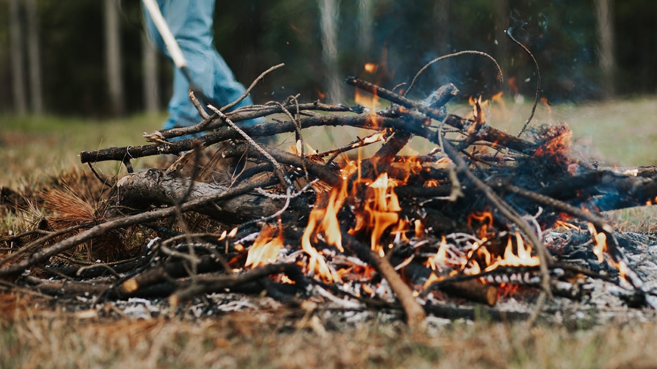 debrisburn-119_websize Low-angle view of a large bonfire burning brightly in an outdoor grassy area. The fire consists of dry branches and logs, with dynamic orange and yellow flames, rising grey smoke, and visible ash beneath.