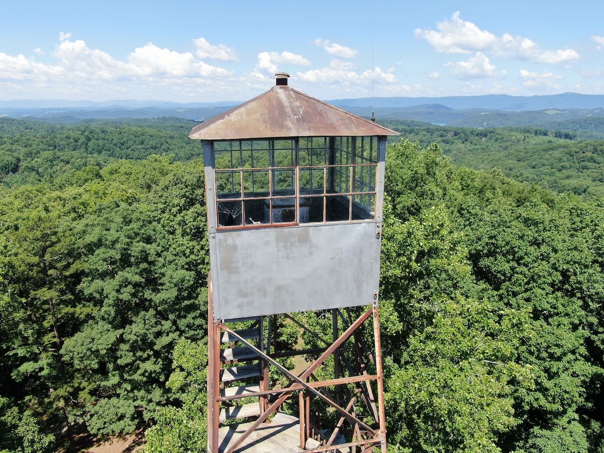 Chuck Swan State Forest 2023 2 A solitary, weathered fire lookout tower with a rusted metal roof and visible staircase, surrounded by a vast green forest under a blue sky with scattered white clouds, emphasizing the tower's historical significance and isolation.