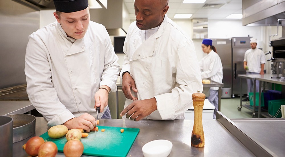 Apprenticeship TN A chef supervisor watches over a young chef cutting vegetables