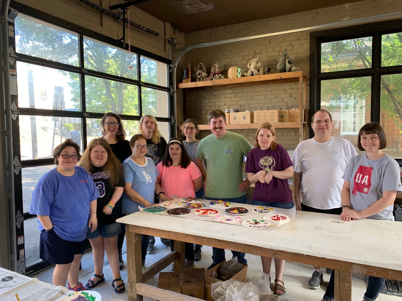 Group photo of seven DAART participants from Carroll County, two Teaching Artists, Sarah Edwards and Candace Gooch, and two volunteers, Arianna Whaley and Sel Montgomery a group of adults with and without disabilities standing behind a table with art supplies posing for a photo