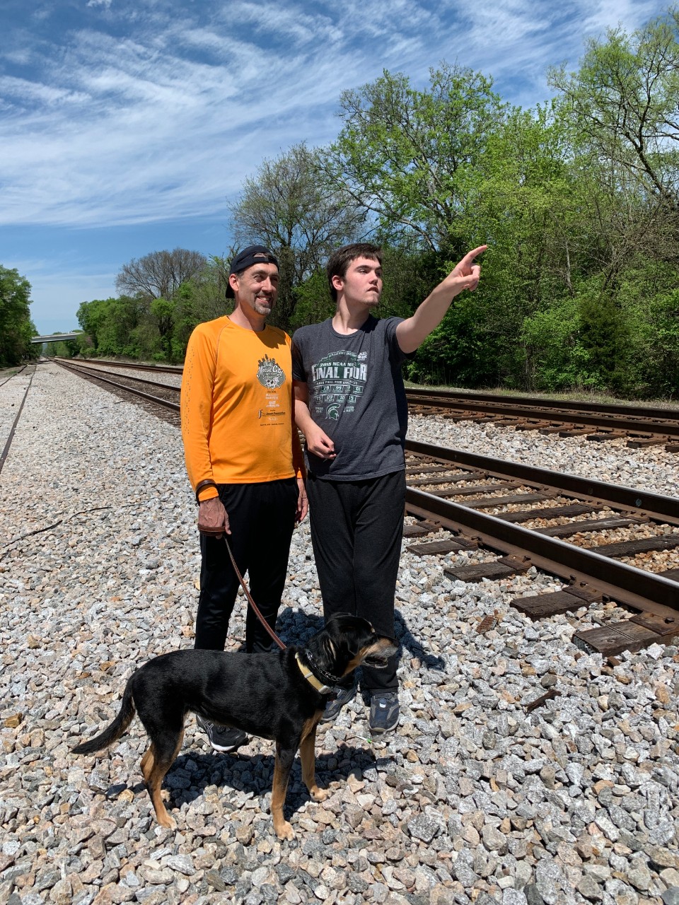 A go-to activity with Aidan is to walk to the train tracks A photo with Aidan and his father standing next to train tracks on a beautiful sunny day, and Aidan pointing to a train past the camera. They're also walking their medium sized black dog.