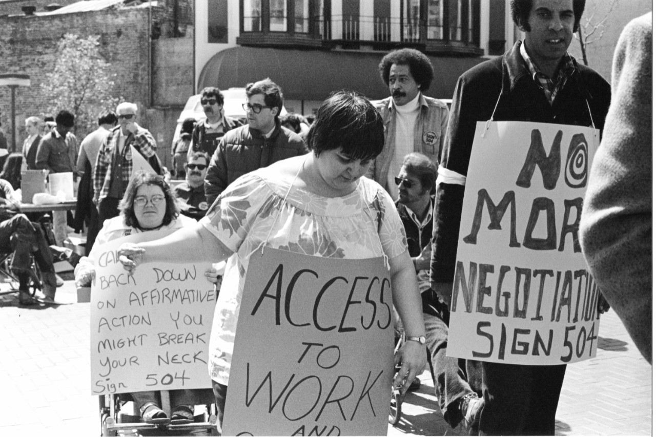 Protesters at UN Plaza outside the HEW Building, April 5, 1977 black and white photo of a diverse group of people, Black and white and with and without visible disabilities marching. A few protest signs are partially visible. One says “No More Negotiation – sign 504.” Another says “Access to work.” Several of the protesters are people using wheelchairs.