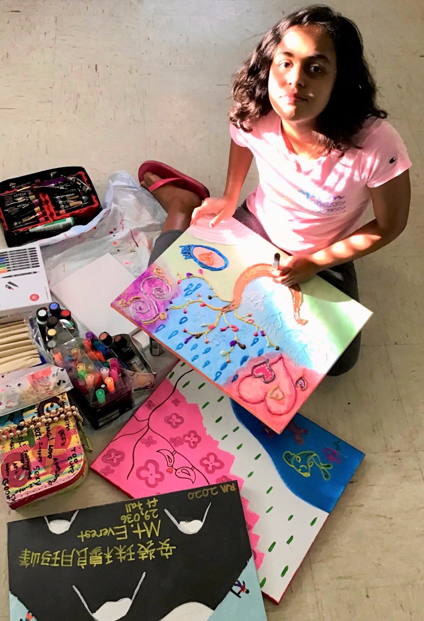 Rainbow Mosho at work with art supplies purchased through the Council grant photo of the young artist – she is shown sitting on the floor with several paintings of hers around her and in her lap, doing her art. There’s sun from a window shining on her face, and she has a hint of a smile