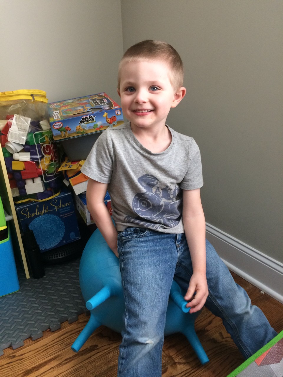 Corban Neale a smiling young blonde boy in a gray t-shirt, sitting on the blue yoga ball with toys and puzzles behind him