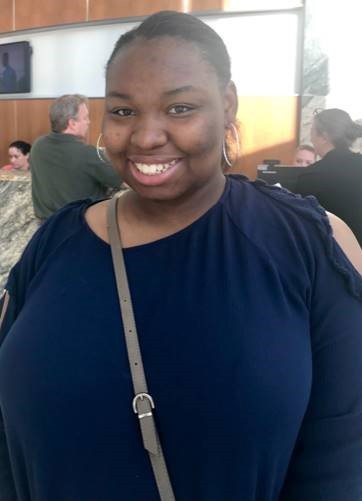 L’Mya is a young Black woman and the small photo of her shows her smiling, wearing a black shirt and she has her hair pulled back.