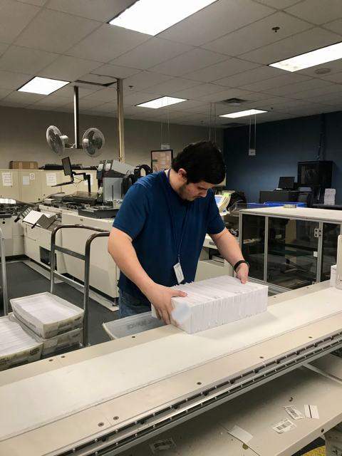 Manases is a young man shown in a large mailroom by himself, concentrating on holding a big stack of mail to transfer it to another box. 
