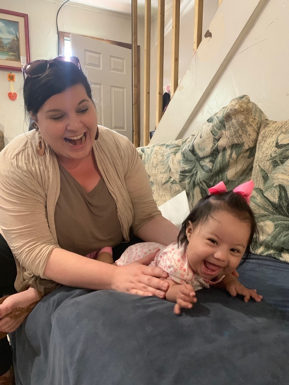 In a later teletherapy photo, Sofia is active and engaged in her therapy visit, showing her great progress. The picture shows a happy Latino infant girl laying on her belly on a couch, being held by her happy and smiling mom, as Sofia wiggles her arms and legs.