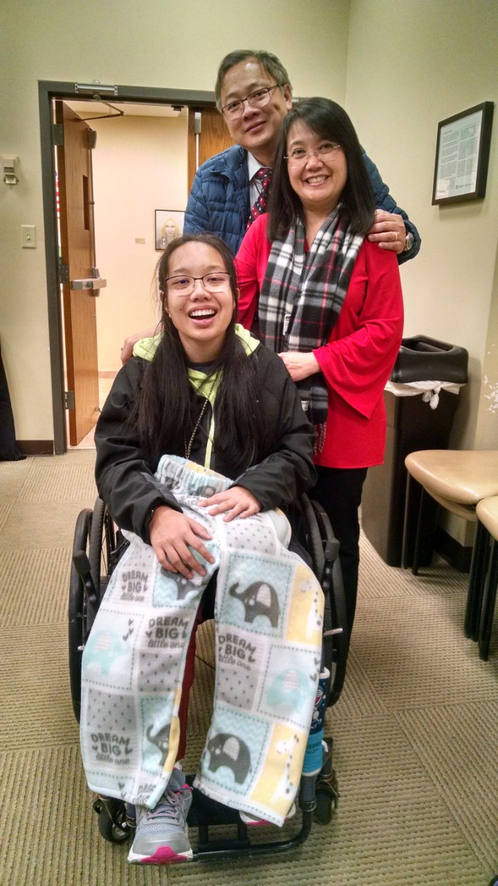 Peach adaptive fashion A young Asian woman with long black hair, glasses and a smile sits in her wheelchair with her parents standing behind her. She’s holding soft fleece patterned pants across her legs.