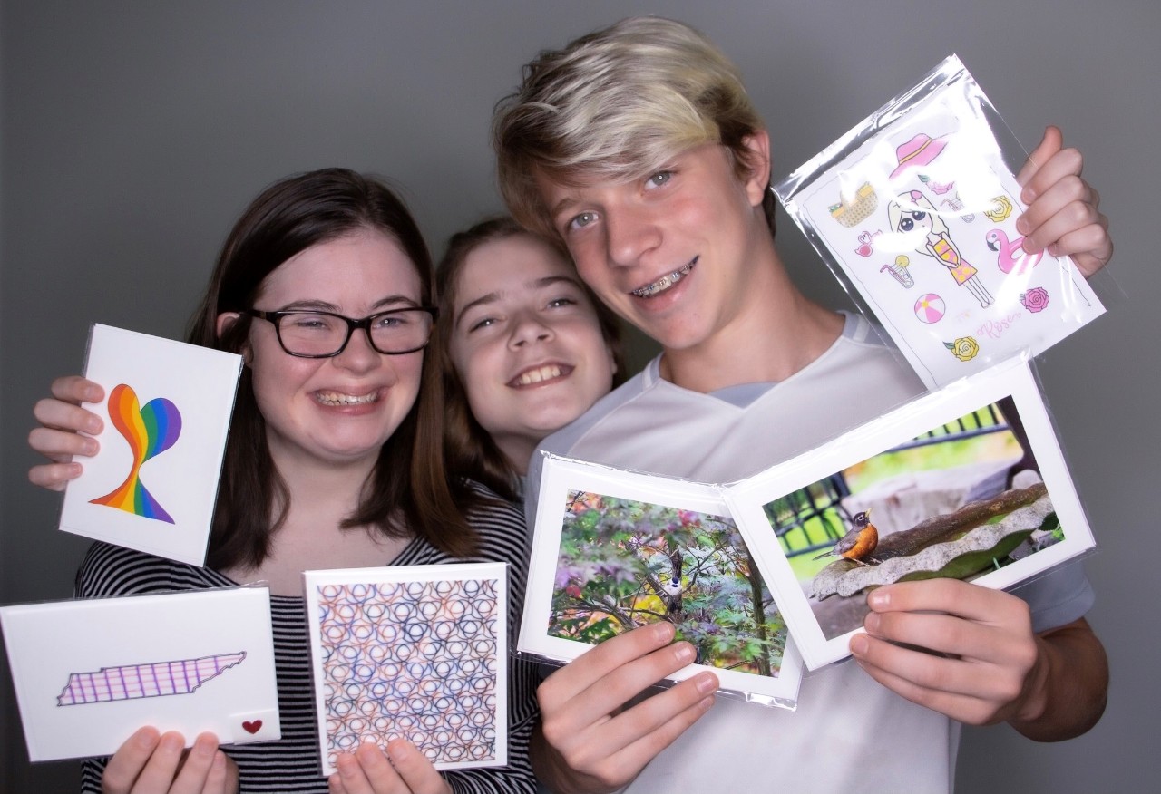 Marie, Elizabeth, and Thomas Tisdale showing their cards for Jessamine Studios. The photo of the three siblings show Marie, a young girl and a teen boy all standing huddled close together with big smiles. Each of the three are holding up multiple colorful notecards that they designed
