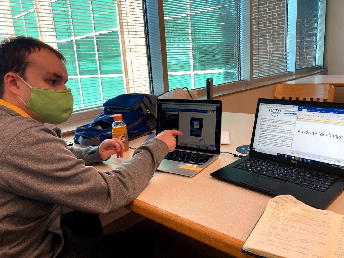 Carter Poland, a young white man wearing a green face mask, seated at a table with a laptop in front of him, working