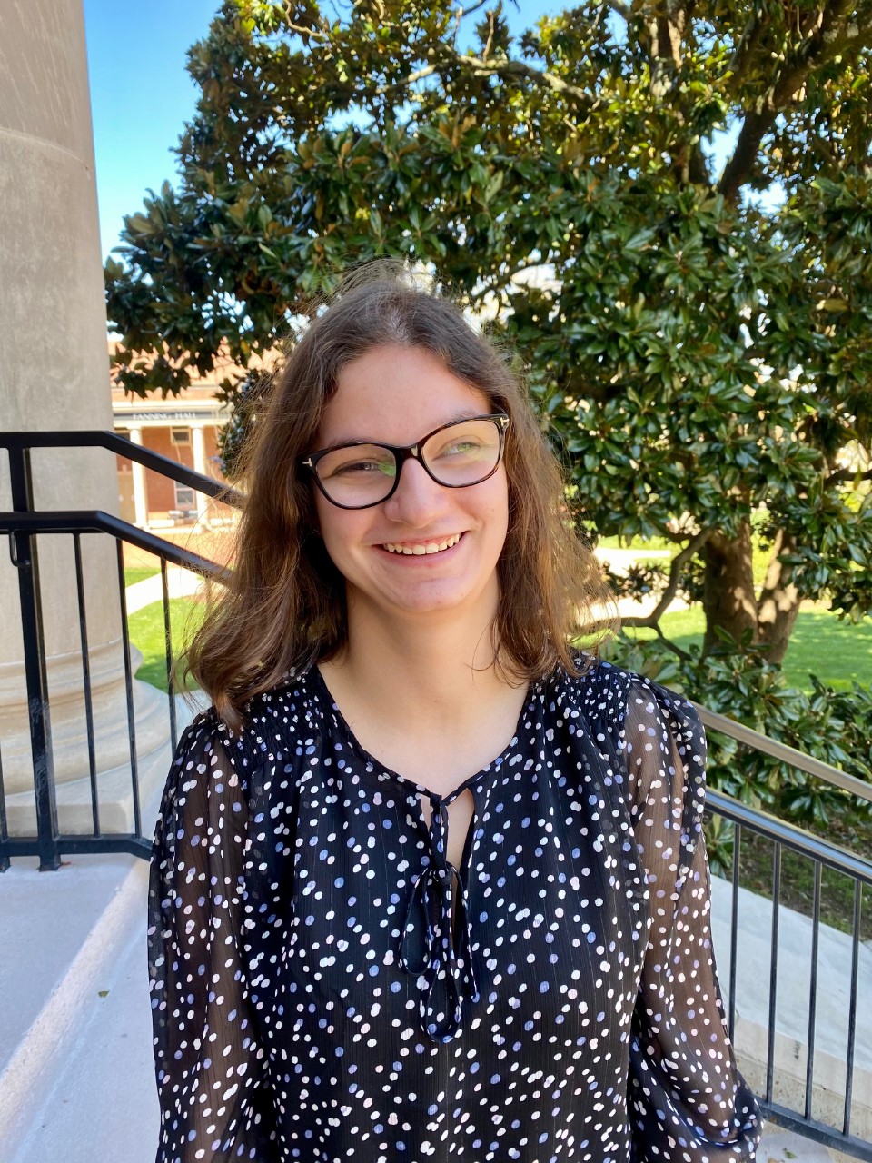 Lauren, a young white woman with glasses and a visual difference, short dark blonde hair and a flowered blouse outdoors at Lipscomb, smiling