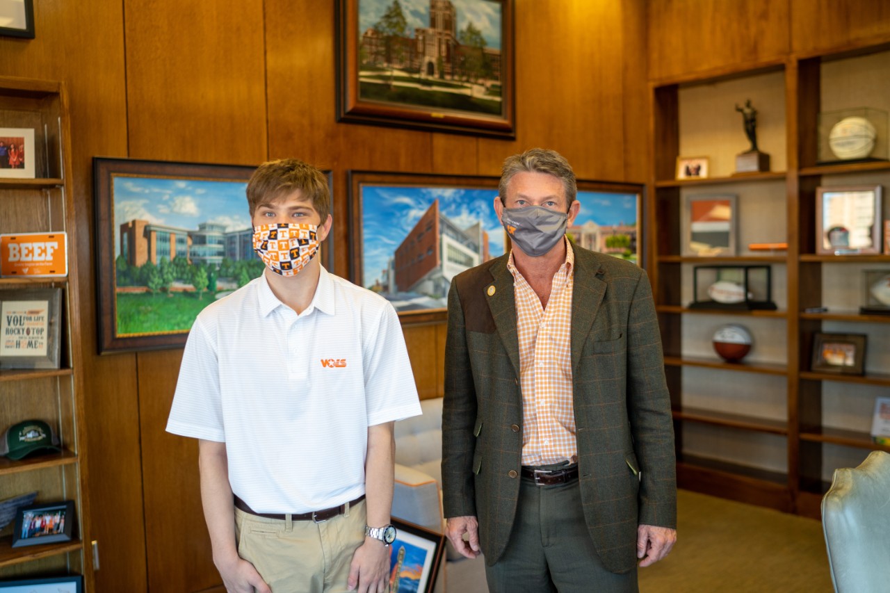Randy Boyd with president’s office intern Coulter Mr. Boyd, UT President, standing in his office wearing a suit, with a young man in a white collared shirt with the UT Vols logo, and UT Vols mask, and khakis.