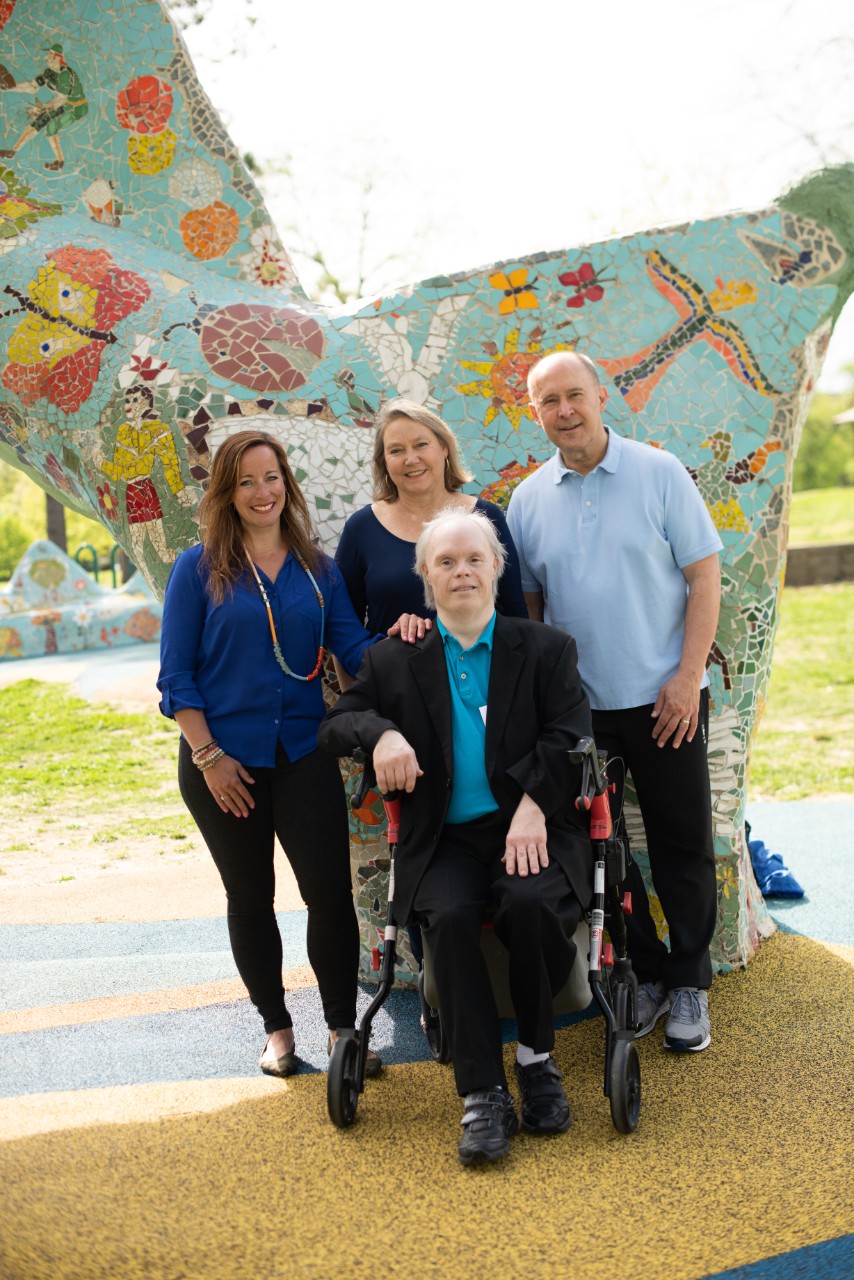 Scott Finney's support circle Photo shows Scott, an older gentleman with Down syndrome with white hair seated on a walker, wearing a nice black suit with a blue collared shirt. Behind him stand his supporters, mentioned in the caption. All are smiling, in front of a blue mosaic statue or mural with pretty insect and flower designs.