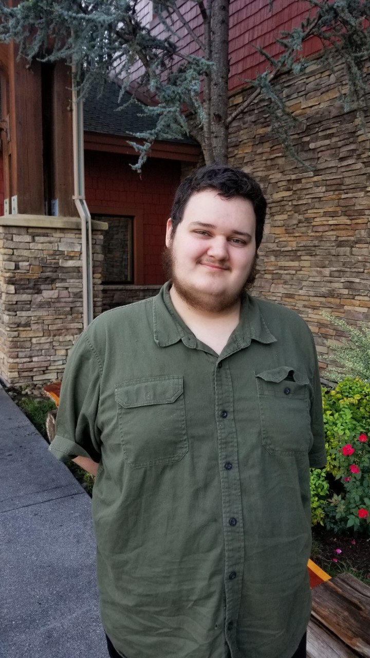 a casual headshot of Mikey Newton, a young adult white man in a dark green button down short-sleeved shirt, with brown hair and a brown beard. He is standing with his hands behind his back and smiling