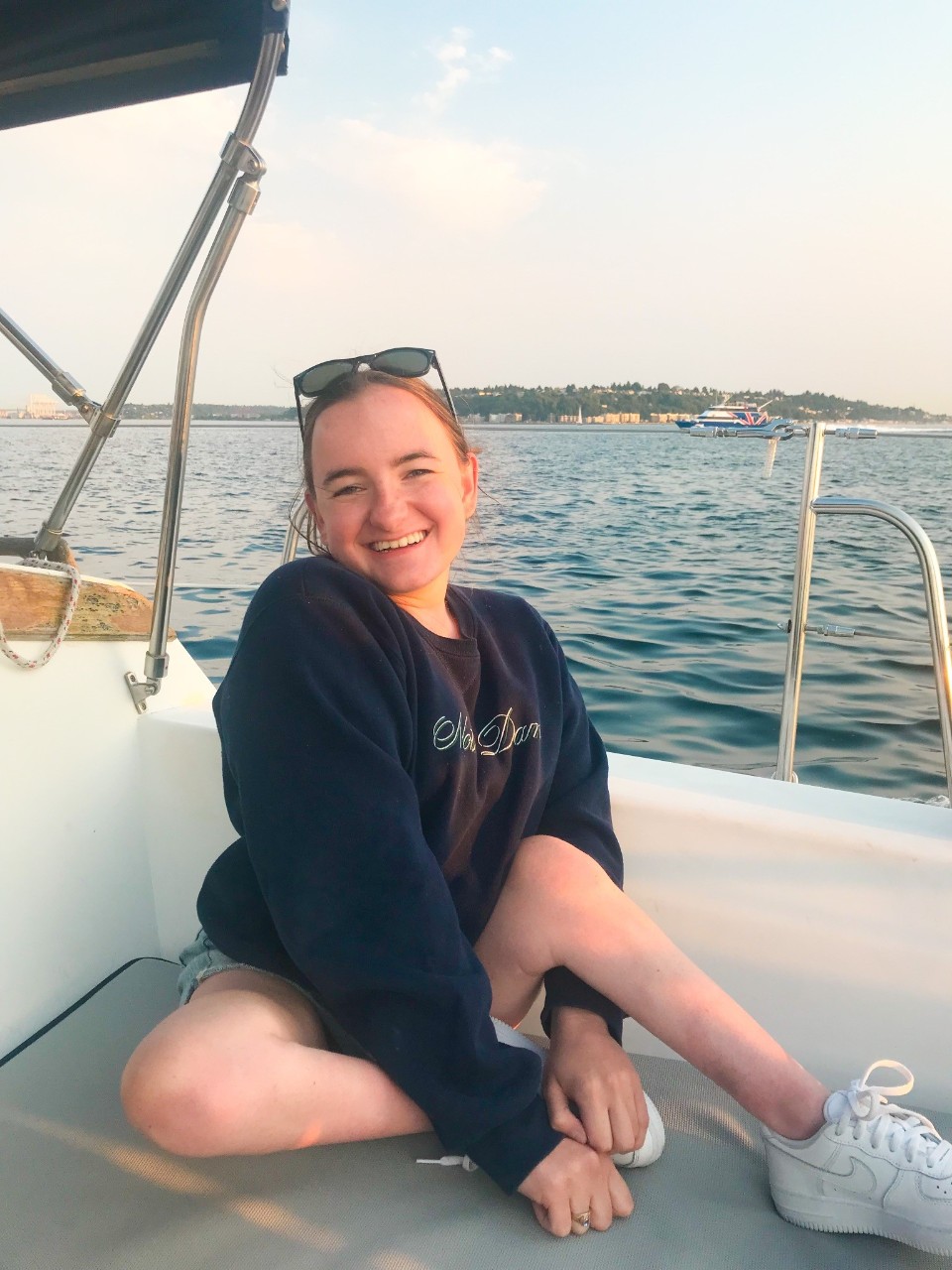 Rosie took a sailing excursion in Seattle, Washington. Rosie took a sailing excursion in Seattle, Washington. She is shown sitting on the bench of a boat, smiling with the ocean behind her. She is in a navy sweatshirt, has her hair pulled back into a ponytail, is wearing sunglasses on the top of her head, and shorts with white sneakers.
