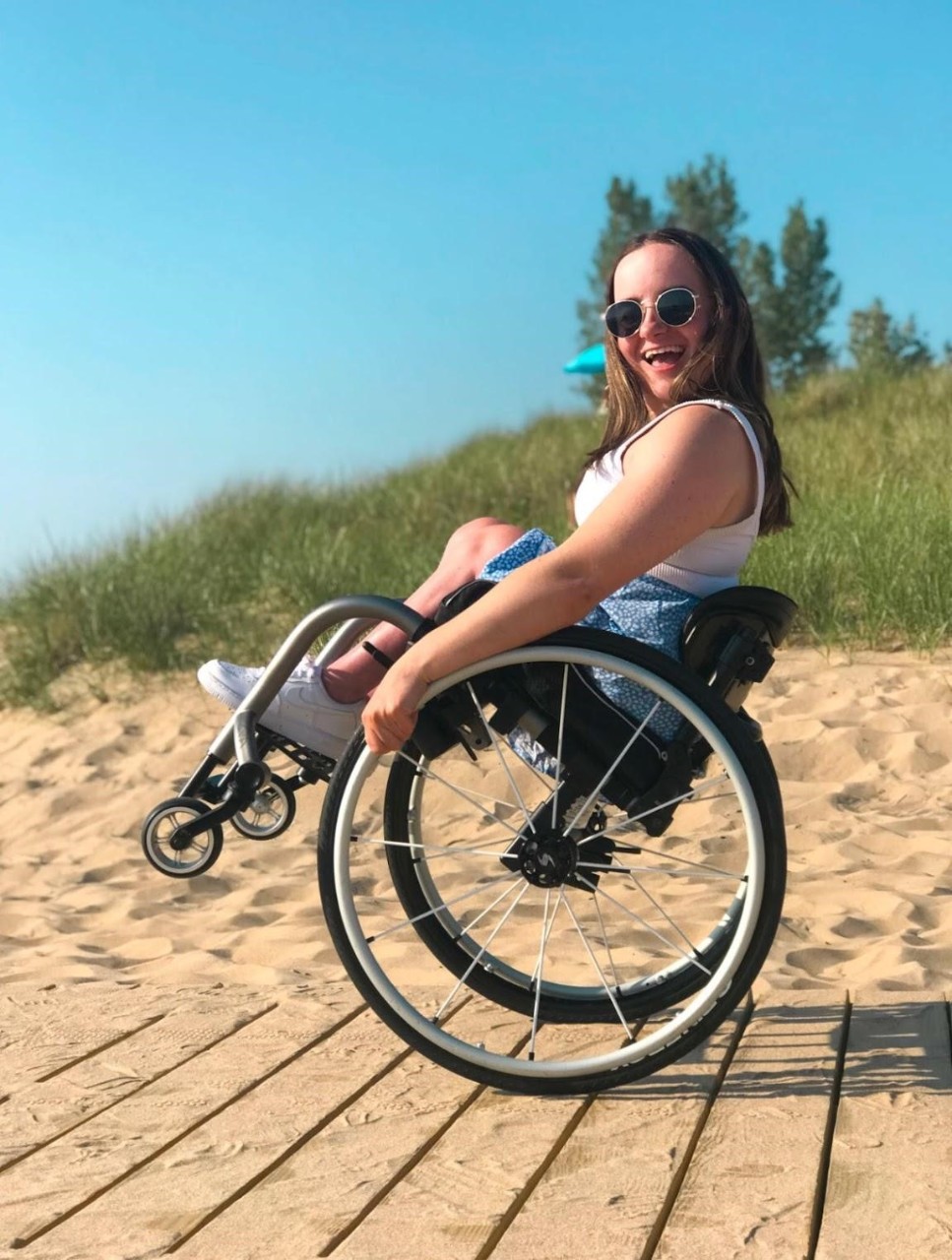 Rosie enjoys the boardwalk at Oval Beach in Saugatuck, Michigan. a photo of a young white woman with a physical disability on a sunny beach that goes with the feature story whose title is also on the cover, “Rosie Roaming: Have Wheels, Will Travel”. She has long brown hair, a white tank top, blue flowered shorts, white sneakers and sunglasses. She is tilting back doing a wheelie in her wheelchair and smiling a big smile. She is on a wooden boardwalk on a beach with sand and beach grass behind her. It is a beautiful sunny day with blue skies.