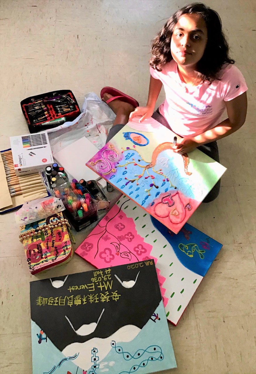 2020 COVID grants - Rainbow Mosho artwork A young girl with dark skin and dark, wavy hair looks up from her seated position on the floor, surrounded by paintings and art supplies. Slatted sunlight shines on her face and pink t-shirt