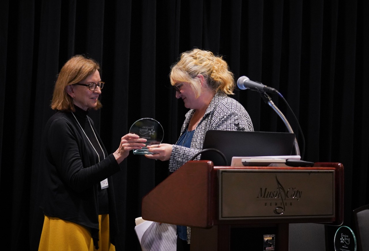 Wanda receiving DRT Freedom Award 2018 Wanda, wearing a black top and gold-colored skirt, accepts a round, glass award from Lisa Primm, a white woman with wavy blond hair and glasses, wearing a blue geometric blazer over a blue top. A large podium in the foreground, and the background is black curtains.