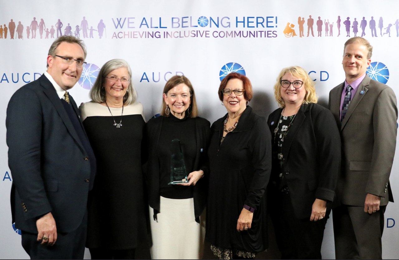 2018 AUCD Andy Imparato with TN DD Network Six people in dark dress clothes pose in front of an event background. The banner says at the top, “We all belong here! Achieving Inclusive Communities.” Wanda is in the center of the group, with a white man and a white woman with gray hair on the left, and two white women and a white man on her left. Wanda is holding a clear award