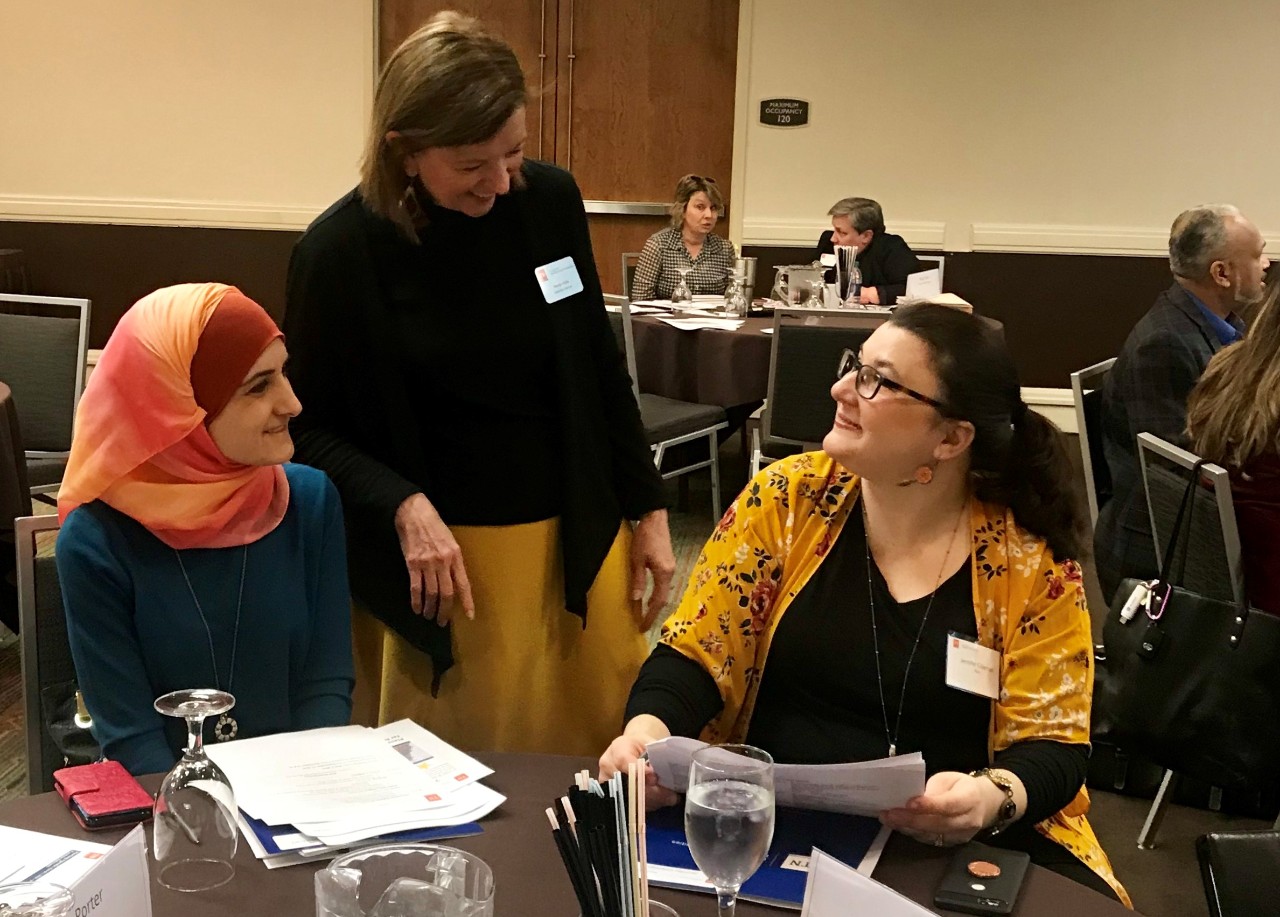 Wanda chats at a 2019 Council meeting with members Sarah Kassas and Jennifer Coleman. A photo of Wanda standing between two women talking to them as they sit at a table in a meeting room