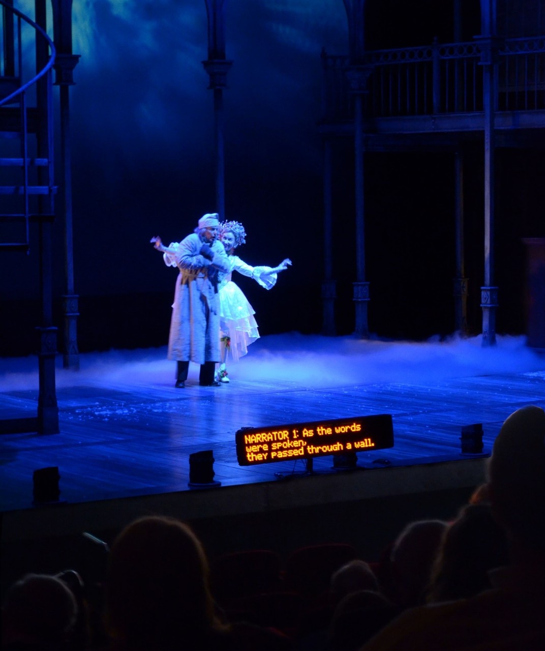 David Kortemeier and Katy Wolfe, in Clarence Brown Theatre’s production of A Christmas Carol. Captions are visible at the front of the stage. Photo by Natasha Koetsch two actors on a theater stage, with electronic captions being displayed to the audience at the foot of the stage