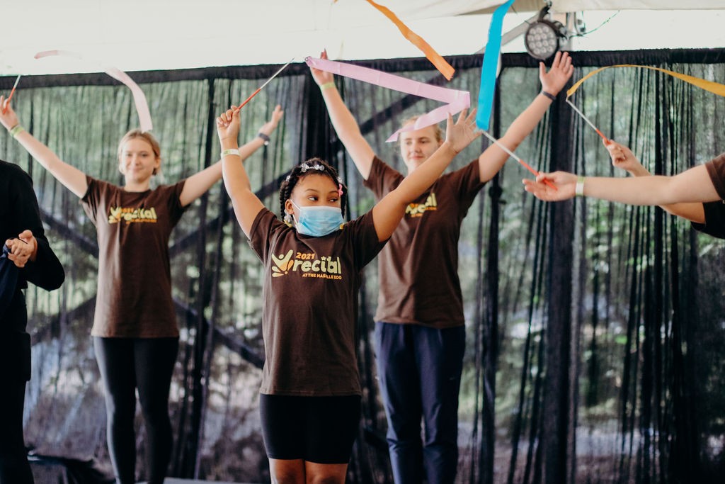 Dance student Saniya Rose (front) and volunteers perform a ribbon dance at a 2021 Recital at the Zoo a young Black girl is on a stage with a few other dancers behind her, and all of the dancers are waving colorful ribbons on sticks in the air with their arms raised. They are all wearing matching shirts with the “MOVE” dance program logo that say “2021 Recital”.