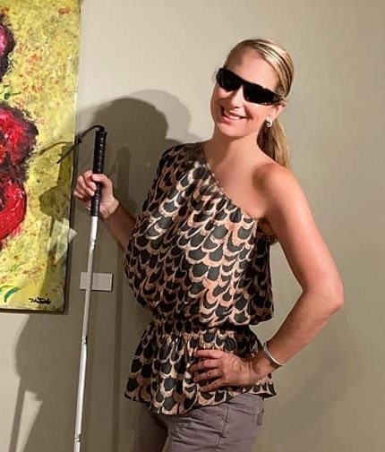 Tracy Bettencourt, owner/artist of Bettencourt Originals 1. Photo of Tracy shows her posing beside one of her painting in a gallery and smiling with her hand on her hip. She is a white woman with long blonde hair pulled back in a ponytail wearing dark sunglasses, a black and gold patterned blouse that reveals one shoulder, and holding her white cane beside her.