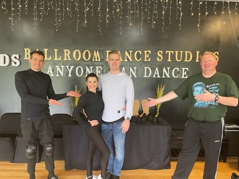Students Ben Rubbelke (far left) and Dave Griffin (far right) celebrate studio owners Brooke Mortimer and Devan McClish. two dance students, Ben and Dave, holding their hands out to present their two dance teachers at the center; all are wearing athletic dancewear and the wall behind them reads “Ballroom Dance Studios – anyone can dance.”