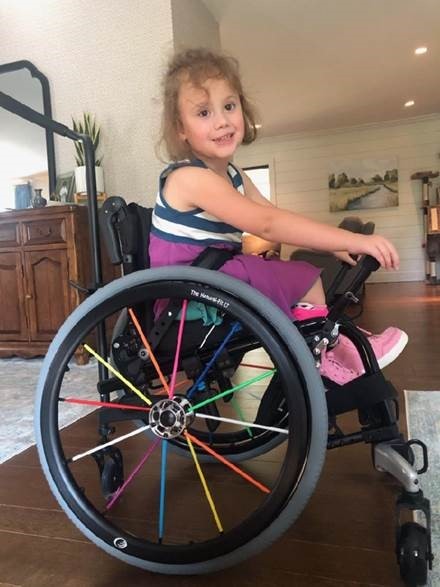 Katie Beckett paid for a Freewheel accessory so Emory can take her wheelchair off-road to join in the fun with family and friends a young little girl with curly hair in her wheelchair in her house, smiling.