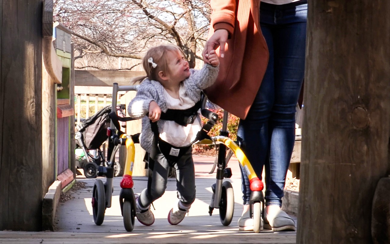 Two-year-old Cosette Smith loves to be on the move. Her walker was purchased with the support of Katie Beckett Part B a very young little girl suspended by a walker, shown holding her mom’s hand as they walk together down the sidewalk. She is looking up at her mother and smiling.