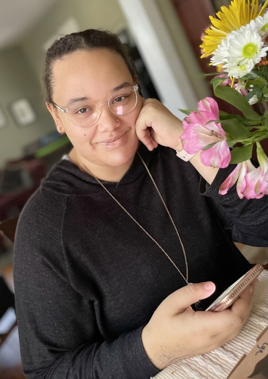 Amyra Perkins young woman of color sitting at a desk in a black hoodie with her hair pulled back and a nose stud and clear rimmed glasses, with a bouquet of flowers