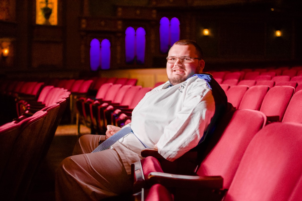 Jacob Collins, also known as Super Jacob young white man sitting in red velvet seats in an empty theater; wearing glasses and a blue cape