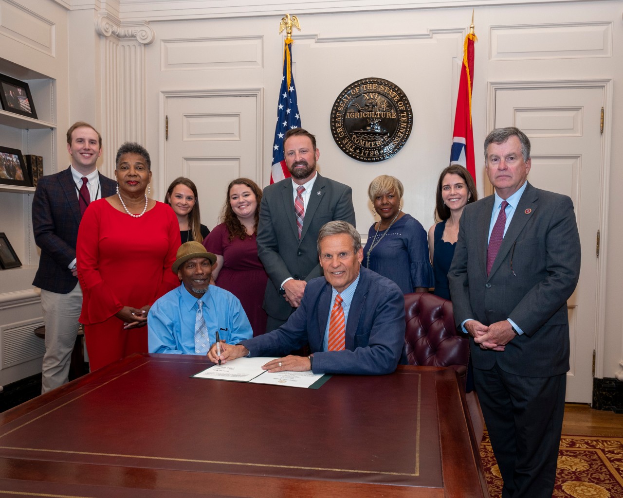 group of about a dozen people seated and standing by the Governor at the capitol while he signs the legislation, posing for a group photo