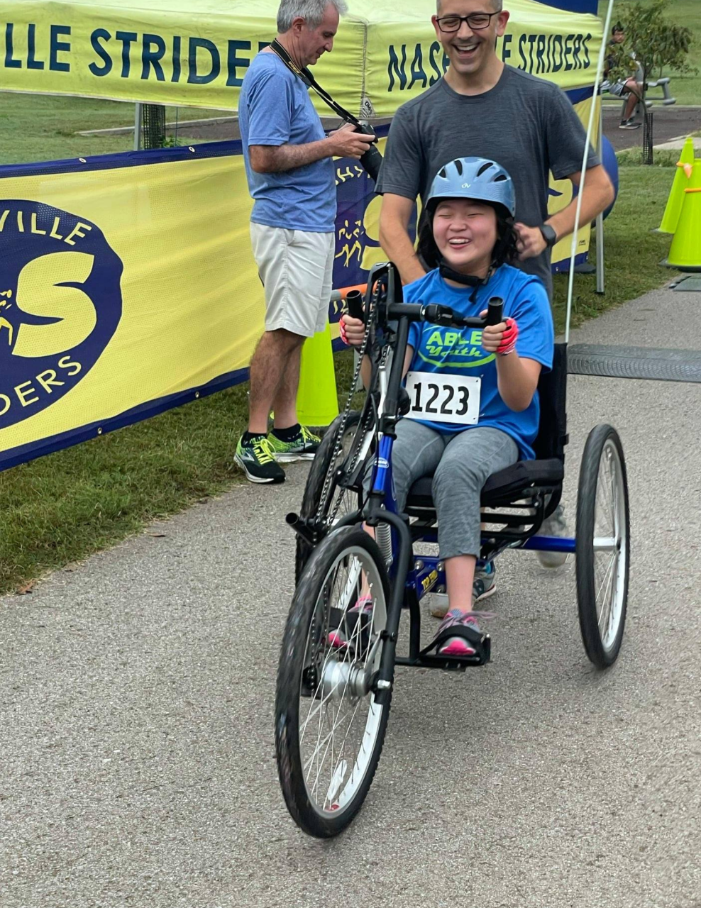 a girl is seated on a large accessible tricycle that is low to the ground and an adult is running along behind her, helping push during the race