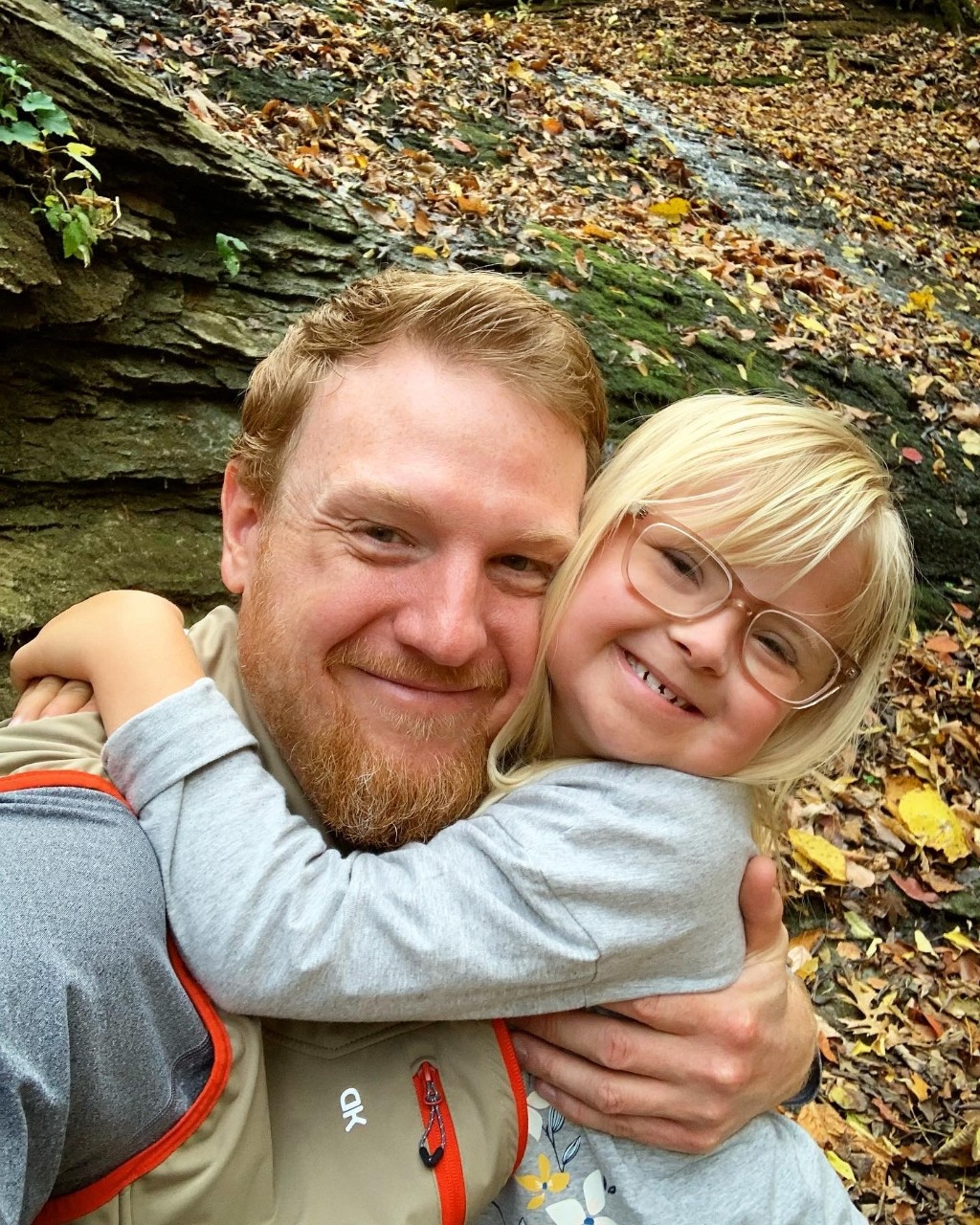 Jon and Lina Sharp Jon is a young dad with reddish short hair and a beard and Lina is a young girl with light blonde hair and glasses who is tightly hugging her dad’s neck with a big grin on her face, fall leaves in the forest behind them