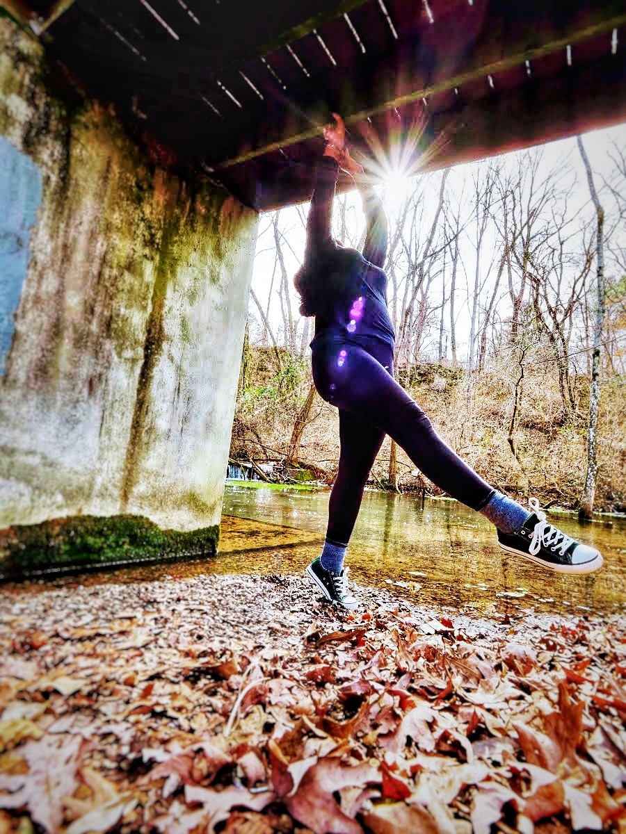 Jennifer James a young black woman shown in a dance pose, leaping in the air with one leg kicked out in front of her and her head back and arms up; she appears to be under a bridge with leaves covering the ground under her feet, and a creek, forest and shining sun behind her