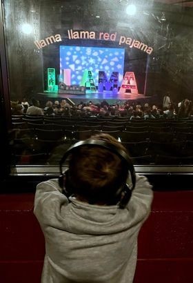 photo at the Nashville Children's Theater with "Llama Llama Red Pajama" set design on stage, with the a child wearing noise canceling headphones leaning forward and watching the play from their new sensory room