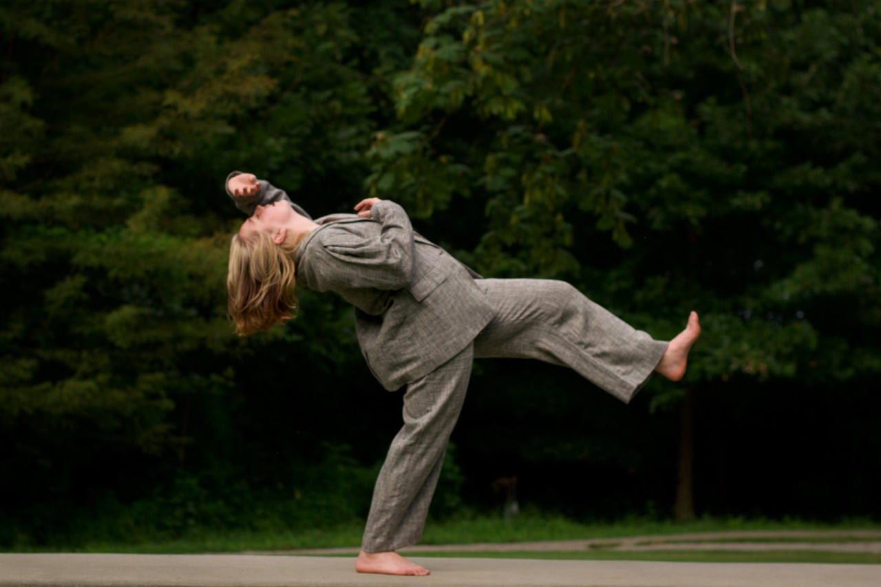 photo of a person with medium length blonde hair dressed in a baggy light gray suit and jacket in a dramatic pose in profile in front of a forest. The person is balancing on one foot with the other extended up in front of them and their body leaning backwards, with one arm draped above their chest and the other with their hand on their forehead. 