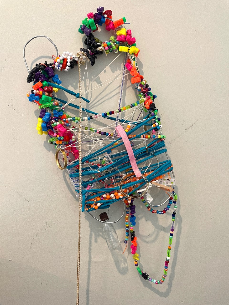 mixed media piece formed from bent wire and colorful beads and pom-poms and ribbons and string wrapped around it; shape is sort of like a bottle being poured out upside down