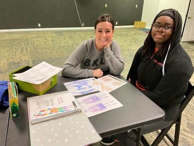 Backlight Productions Executive Director Melissa Smith and Danielle Agholor discussing and sketching costumes two women sit at a table together that is covered in colorful sketches of costumes and smile for the camera
