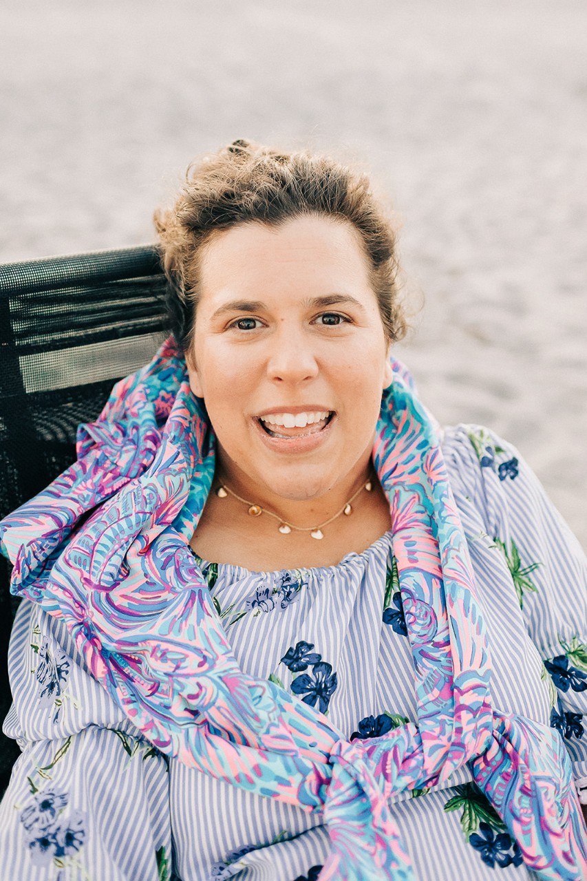 Samantha Siedlecki, President & Founder of Paradise Shells and Fine Jewelry by Samantha a portrait of a young white woman with short dark blonde hair sitting in her wheelchair on a sandy beach, wearing a colorful scarf and blue flowered blouse and a pretty silver necklace