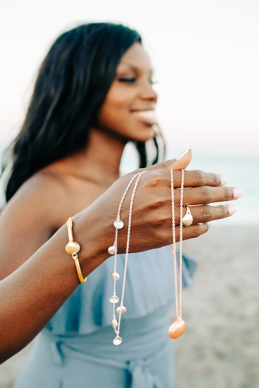 A model shows off several Paradise Shells items. closeup photo of a black female model standing on a beach, with silver and gold necklaces, bracelet and ring draped over her outstretched hand and forearm