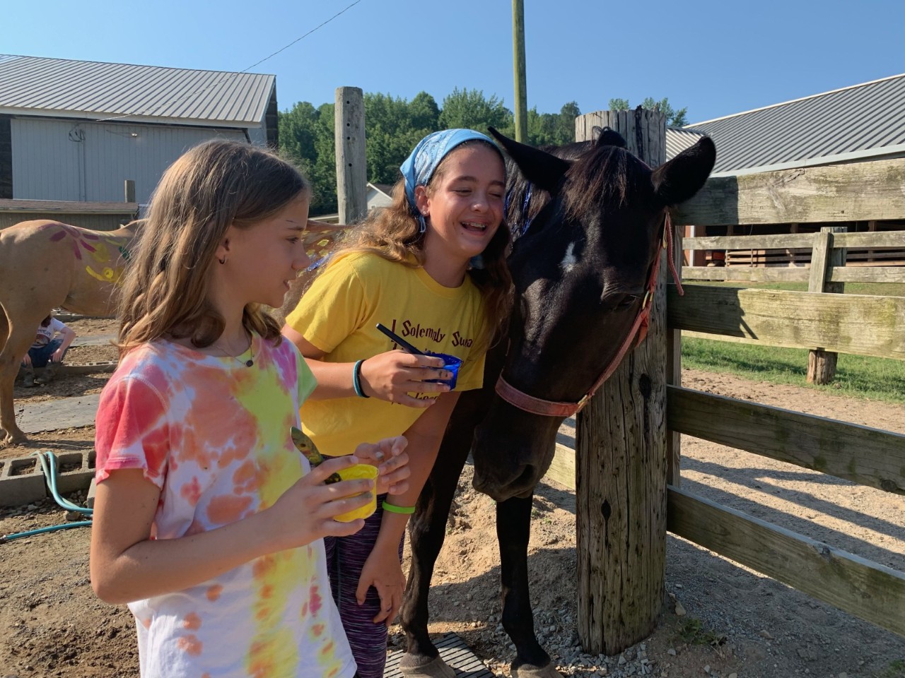 two young girls lean near a black horse’s head at an outdoor fence while laughing; there’s a brown horse in the background with painted handprints on its side