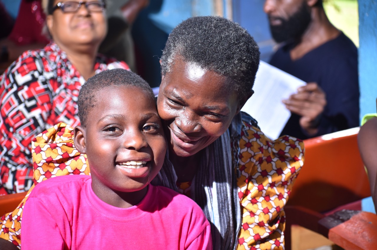 A parent event offered a sense of hope and community to families who were gathering together for the first time a young child in a bright pink shirt and an adult female family member with a colorful dress is shown leaning over his shoulder - the boy is looking at the camera directly and smiling, his family member is smiling down past him