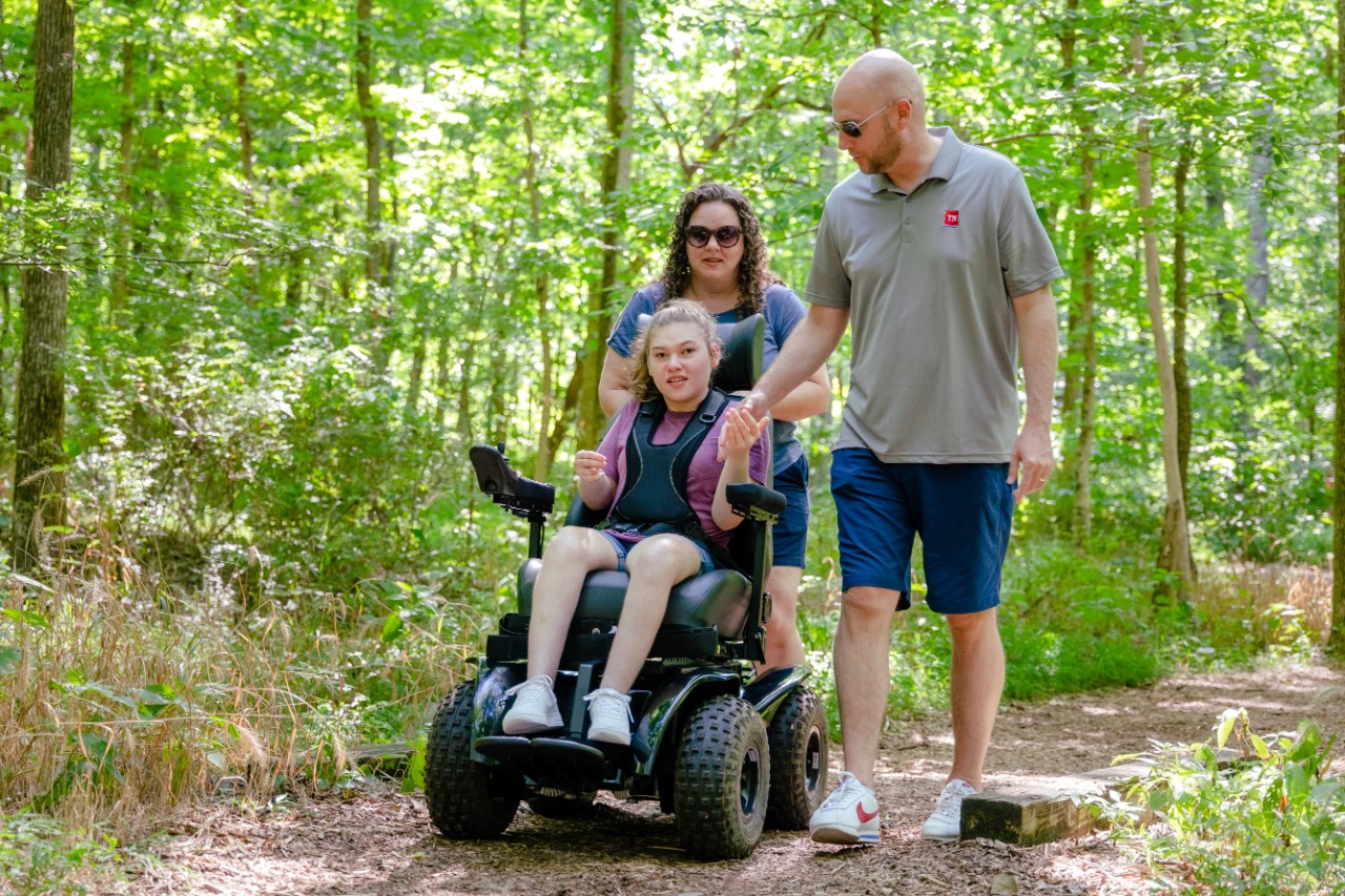 Turner family photo with all-terrain wheelchair DIDD Commissioner Brad Turner, a white man with athletic clothes on, stands next to his wife Rebecca on a nature trail at Radnor Lake park with their daughter Kinsley, a teenage wheelchair user, in an all terrain wheelchair
