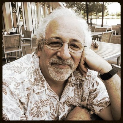 A sepia-toned photo shows a white man with gray hair and mustache and goatee smiling at the camera, his head resting against his left hand. He is wearing a Hawaiian-style collared shirt, glasses, and a dark colored watch. 
