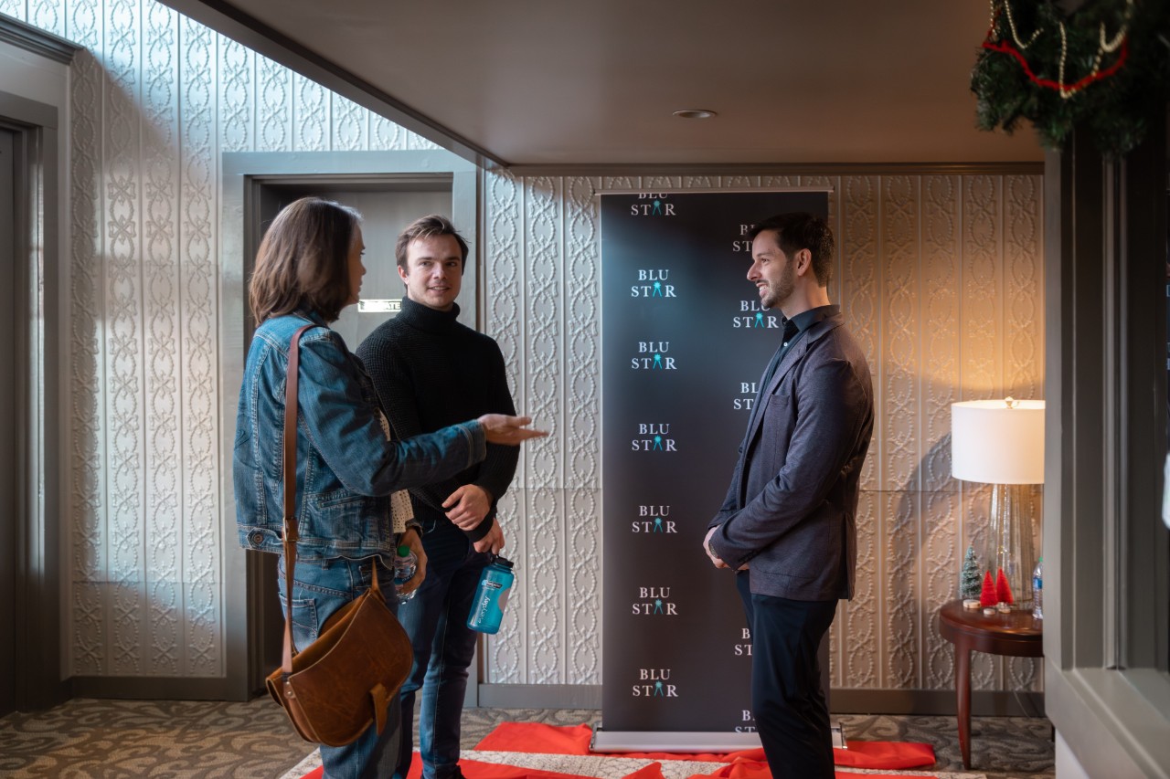 A young white man in a brown suit jacket and slacks listens as a couple of people talk to him, and all are standing in front of a photo backdrop with the Blu Star logo on it.