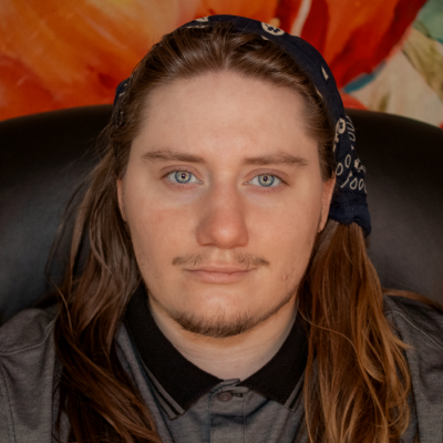 Dirk Strider Image Description: A close-up portrait of a light-skinned person with long brown hair and blue eyes. He has a slight mustache and is wearing a dark-colored bandana with a pattern, tied around his head. He is also wearing a gray shirt with a black collar. The background is colorful, with warm tones and abstract shapes. The person is looking directly at the camera with a neutral expression.